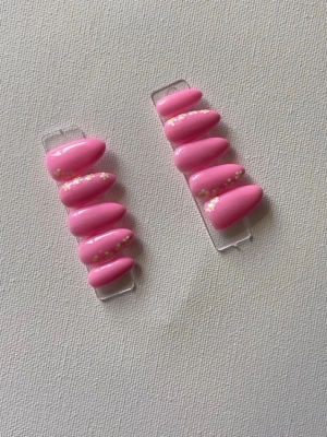 Rosa press-on naglar med blommor - Säljer ett set press-on naglar i en snygg rosa färg med mandelform. Några av naglarna har små vita och gula blommor som dekoration. Perfekt för dig som vill ha en söt och trendig look. 