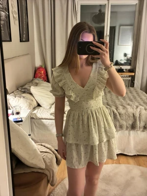 Ljus kortklänning med volang och v-ringning - Söt ljus kortklänning med små blommönster, v-ringad och volangdetaljer vid axlar och kjol. Klänningen har puffiga kortärmar och smockad midja som ger en feminin siluett. Perfekt för sommarens alla tillfällen.