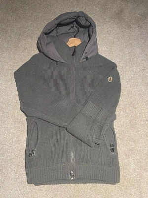 Grå Moncler cardiganjacka med huva - Snygg grå cardiganjacka från Moncler med stickade ärmar och framstycke samt quiltad baksida och huva. Jackan har dragkedjor på fickorna och en broderad Moncler-logga på ärmen. Perfekt för lager-på-lager och streetstyle. De går o diskutera om priset vid snabbäffar. 