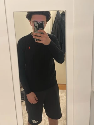 Svart stickad tröja från Polo Ralph Lauren - Säljer en svart stickad tröja från Polo Ralph Lauren med klassiskt kabelstickat mönster och röd logotyp på bröstet. Tröjan har rund hals och långa ärmar, perfekt för att hålla dig varm och snygg under kalla dagar. Är storlek L men passar S och M 