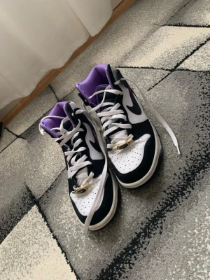 Nike Dunk High svart/vit/lila sneakers - Säljer ett par Nike Dunk High sneakers i svart och vitt med lila innerfoder. Klassisk Swoosh på sidan, vita skosnören och guldfärgade dubraes. Ovandelen är i läder och sulan är vit med svart yttersula. Perfekta för dig som vill sticka ut med coola färgdetaljer. Skorna har ett bra skick 