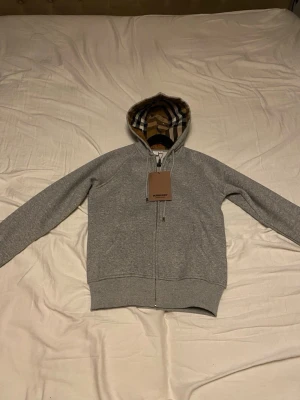 Grå zip hoodie Burberry - Snygg grå zip hoodie Burberry med klassiskt rutigt mönster i huvan. Tillverkad i mjukt material och har dragsko i huvan samt ribbade muddar vid ärmar och nederkant. Perfekt för en chill och stilren look.
