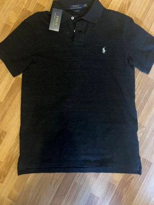 Svart piké Ralph Lauren - Klassisk svart pikétröja från Polo Ralph Lauren i slim fit-modell. Tröjan har korta ärmar, krage med knappar och den ikoniska loggan broderad på bröstet. Perfekt till jeans eller shorts för en clean och stilren look.