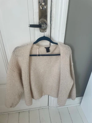 Beige stickad kofta från Lindex - Mysig beige stickad kofta från Lindex med öppen front och avslappnad passform. Koftan har breda ärmar och är gjord i ett mjukt, fluffigt material som känns skönt mot huden. Perfekt att slänga över en t-shirt eller topp för extra värme.