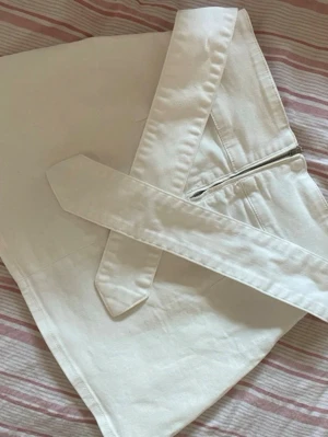 Vit kortkjol med bälte och dragkedja - Säljer en stilren vit kortkjol med tillhörande bälte och synlig dragkedja i sidan. Kjolen har en enkel, rak passform och är gjord i ett mjukt bomullsmaterial. Perfekt för dig som gillar minimalistisk och clean stil.