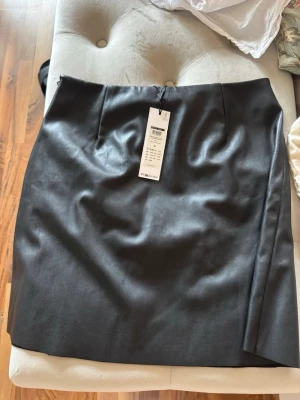 Svart skinnimitation kjol från Noisy may  - Snygg svart kjol i skinnimitation från Noisy may. Kjolen har en rak, kort modell med diskreta sömdetaljer framtill och dragkedja bak. 