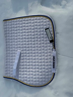 Vitt schabrak Hansbo Sport Ponny - Säljer ett vitt schabrak från Hansbo Sport i Ponny-storlek. Schabraket är vitt med blå och gul kant samt praktiska fästen. Perfekt för dig som rider ponny och vill ha en stilren look till utrustningen.