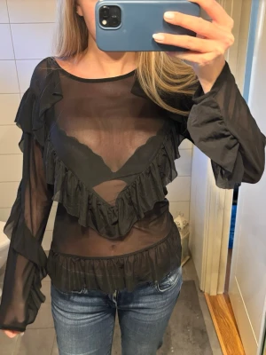 Genomskinlig festtoop - Säljer en svart genomskinlig blus med långa ärmar och snygga volanger framtill och vid midjan. Storlek M från MSCH Copenhagen.