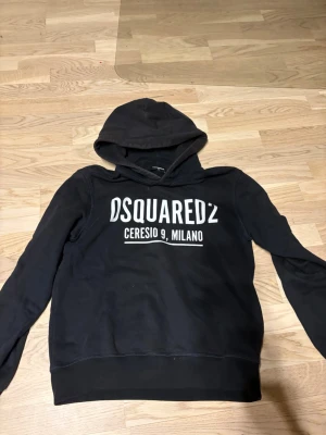 Svart hoodie från Dsquared2 - Svart hoodie från Dsquared2 med stor vit logga och texten 'Ceresio 9, Milano' på bröstet. Klassisk modell med huva och långa ärmar. Tillverkad i mjukt bomullsmaterial som känns skönt mot huden. Perfekt för en avslappnad och trendig look.
