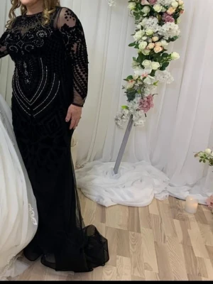 Svart långklänning med glittriga detaljer - Elegant svart långklänning med transparenta ärmar och glittriga paljettdetaljer. Klänningen har en figurnära passform, rund halsringning och vackra mönster framtill. Perfekt för dig som vill sticka ut på festen.