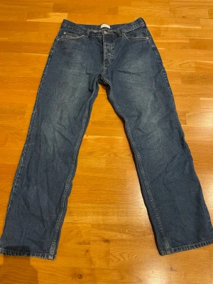 Blå raka jeans i klassisk modell - Ett par blå jeans med rak passform och fem fickor. Jeansen har en klassisk midja och stängs med dragkedja och knapp. Tillverkade i denim med tydliga sömmar och enkel design, perfekta för en avslappnad stil. Stolek 32