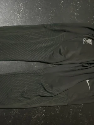 Svarta Nike LFC träningsbyxor - Säljer ett par svarta träningsbyxor från Nike med Liverpool FC-logga på ena benet. Byxorna har diskreta mönster längs benen och är tillverkade i ett lätt, sportigt material som passar perfekt till träning eller fotboll. Det är ett 2 millimeters hål som jag kan sy