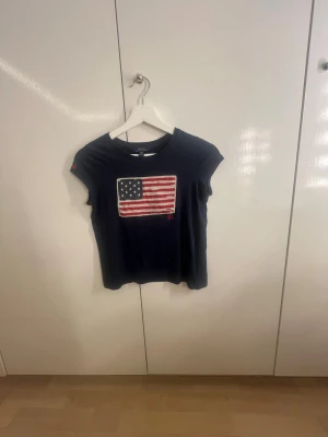 Mörkblå t-shirt med USA-flagga RL - Snygg mörkblå t-shirt från Ralph Lauren med en stor USA-flagga broderad på bröstet och RL-detalj. T-shirten har korta ärmar och en klassisk passform. Perfekt till jeans eller shorts för en avslappnad look.
