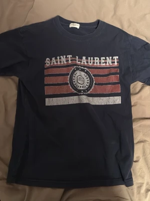 Marinblå Saint Laurent t-shirt - Snygg marinblå t-shirt från Saint Laurent med tryck i vitt och rött framtill. Klassisk passform och rund halsringning. Perfekt för dig som vill ha en stilren men ändå cool look. Tillverkad i mjuk bomull för skön känsla hela dagen.