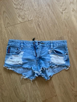 Blå slitna jeansshorts från Levi's - Snygga ljusblå jeansshorts från Levi's med slitna detaljer och fransig kant. Klassisk femficksmodell med låg midja och knappgylf. Perfekta för varma sommardagar och en avslappnad stil.