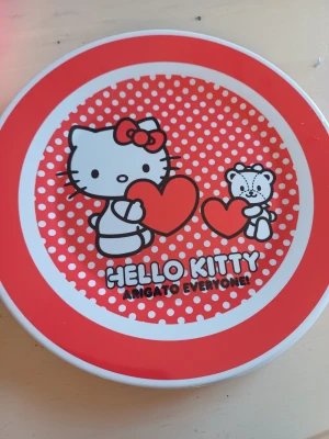 Hello Kitty tallrik med hjärtmotiv - Säljer två  tallrik med Hello Kitty-motiv och texten 'Arigato Everyone!'. Tallriken är vit och röd med prickigt mönster och två hjärtan. Materialet är plast och designen är lekfull och färgstark, perfekt för dig som gillar kawaii och japansk popkultur.