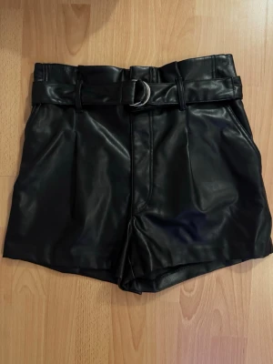 Svarta högmidjade skinnshorts med bälte - Snygga svarta högmidjade shorts i skinnimitation med tillhörande bälte och D-ring. Shortsen har en stilren look med raka ben och markerad midja, perfekta för att lyfta din outfit.