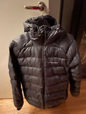 Svart dunjacka från Peak Performance - Snygg svart dunjacka från Peak Performance med huva och dragkedja framtill. Jackan har en quiltad design och logga på bröstet. Perfekt för kalla vinterdagar och riktigt skön att bära. Klassisk modell som funkar till det mesta.
