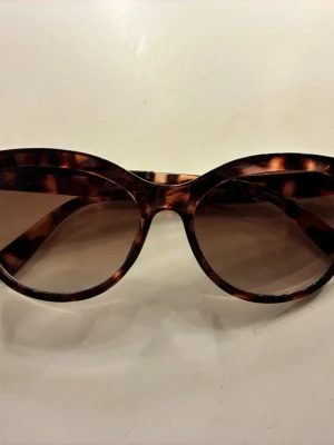 Bruna solglasögon med leopardmönster - Snygga solglasögon med brun leopardmönstrad båge och breda skalmar. Glasen är mörka och har en klassisk rektangulär form som passar till många stilar. Perfekt för dig som vill ha en trendig accessoar med lite attityd.