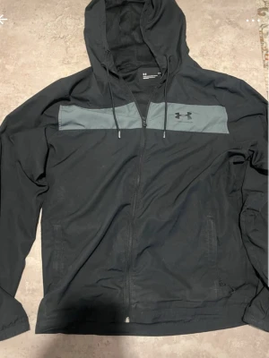 Svart hoodie från Under Armour - Snygg svart hoodie från Under Armour med dragkedja framtill och en grå panel över bröstet. Hoodien har justerbar huva med snören och Under Armour-logga på bröstet. Tillverkad i ett mjukt och lätt material, perfekt för en sportig och avslappnad stil.