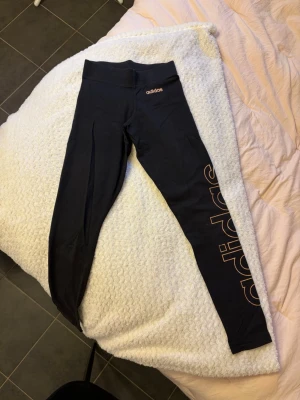 Svarta träningstights från Adidas - Säljer ett par svarta tights från Adidas med stort guldigt logotryck längs ena benet. De har hög midja och är tillverkade i ett stretchigt material som passar perfekt för träning eller chill. Snygg och sportig look med klassisk Adidas-stil. Uppskattning storlek xs