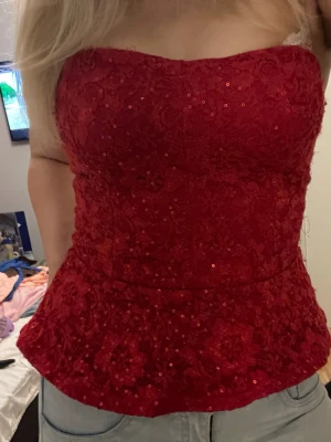 Röd paljettprydd korsettopp - Snygg röd korsettopp med paljetter och spetsdetaljer. Toppen är axelbandslös och har en figurnära passform som framhäver midjan. Perfekt för dig som vill sticka ut med en glittrig och elegant look.