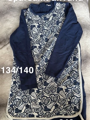 Marinblå långärmad klänning hjärtan - Klänning i marinblått med lång ärm och ett lekfullt mönster av vita hjärtan och glassar på framsidan. Rund halsringning och kontrasterande enfärgade ärmar. Perfekt för dig som gillar unika prints och bekväma plagg.🌸Sparsamt använd. Storlek 134/140. 