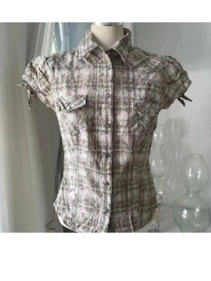 Rutig kortärmad skjorta med rosetter - Kortärmad skjorta med rutig bas i beige och brunt, dekorerad med små rosa blommor. Skjortan har knappar framtill, två bröstfickor och söta rosetter på ärmarna. Perfekt för dig som gillar romantiska detaljer och en avslappnad stil.