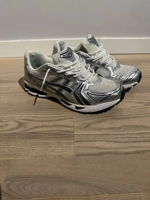 Asics vita och silver sneakers - Snygga sneakers från Asics i vitt och silver med svarta detaljer. Ovandelen är i mesh och syntetmaterial för bra andningsförmåga och komfort. Klassisk låg modell med snörning och dämpad sula som ger skön känsla hela dagen.