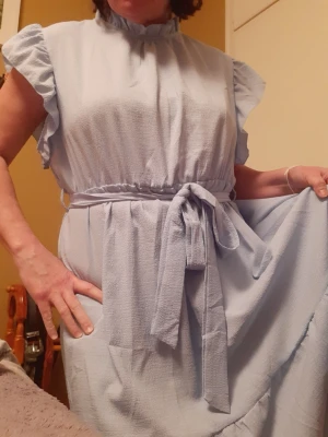 Ljusblå volangklänning med knytband - Söt ljusblå klänning med volangärmar och knytband i midjan. Klänningen har en avslappnad passform och volangdetalj nedtill. Perfekt för sommarens alla tillfällen.