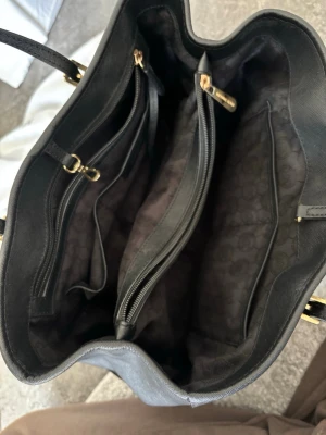 Svart handväska från Michael Kors - Snygg svart handväska från Michael Kors i saffiano-läder med guldfärgade detaljer och logotyp. Väskan har flera fack med dragkedja och innerfoder med diskret mönster. Perfekt för dig som vill ha en stilren och rymlig väska med lyxig känsla.