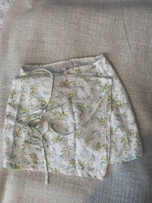 Blommig omlottkjol från Pull&Bear - Superfin kort omlottkjol från Pull&Bear i vitt med blommigt mönster i gult och grönt. Kjolen har knytband i midjan och är gjord i ett lätt och skönt material, perfekt för varma dagar.