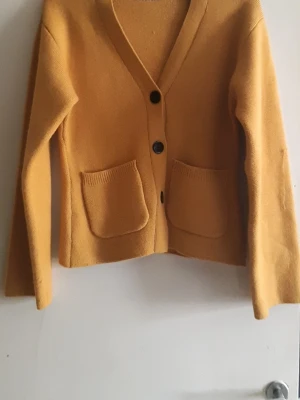 Senapsgul kofta med fickor - Senapsgul kofta med v-ringning och tre svarta knappar framtill. Koftan har två stora fickor på framsidan och är tillverkad i ett mjukt stickat material. Långärmad och enkel design som är lätt att matcha med olika outfits.