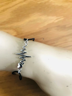 By prisförslag- Svart armband med hjärtslag och hjärta - Snyggt armband med svart justerbart band och ett silverfärgat metallhänge i form av ett hjärtslag och ett litet hjärta. Perfekt accessoar för dig som gillar minimalistisk och modern stil. Passar både till vardagslooken och när du vill addera något extra.