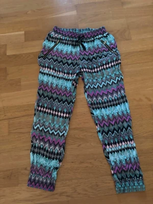 Mönstrade mjukisbyxor från Lindex - Färgglada mjukisbyxor från Lindex med zigzag-mönster i turkos, lila, svart och vitt. Byxorna har resår i midjan och vid bensluten samt sidofickor. Perfekta för en chill och avslappnad stil och sommar.