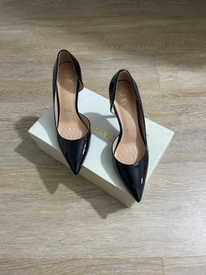 Svarta lackpumps med röd sula - Snygga svarta pumps i lack från  med spetsig tå och hög klack. Skorna har öppen insida och klassisk röd sula som ger en extra touch. Tillverkade i glansigt syntetmaterial och har en elegant siluett som sticker ut.