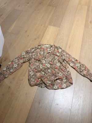Blommig blus från Continue - Blus med brunt botten och färgglatt blommönster i vitt, rött och grönt. Lätt och luftig med krage och knappar framtill. Långärmad modell med smock vid ärmslut. Perfekt för dig som gillar bohemisk och trendig stil.