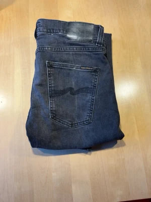 Svarta jeans från Nudie Jeans W31,L30 - Säljer ett par svarta jeans från Nudie Jeans med klassisk femficksdesign och vågig söm på bakfickan. Jeansen har en svart läderpatch i midjan och är tillverkade i mjukt denimtyg. Perfekta för en avslappnad och stilren look.