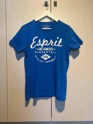 Blå t-shirt från Esprit - Snygg blå t-shirt från Esprit med vit tryckt text på bröstet. Klassisk rund halsringning och korta ärmar. Tillverkad i mjuk bomull som känns skön mot huden. Perfekt för en avslappnad och stilren look.