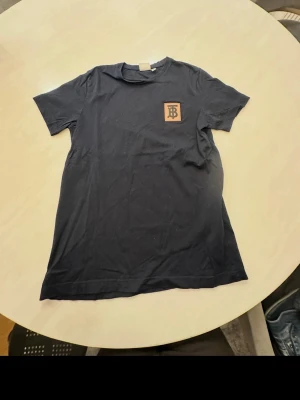 2FÖR1 ) Svart Burberry t-shirt med logga, en mörk blå beige svart logga  - 2flr1 Säljer en svart t-shirt från Burberry med klassisk TB-logga på bröstet. T-shirten har rund hals och korta ärmar, tillverkad i mjuk bomull för en skön känsla. Perfekt för dig som gillar stilrena och exklusiva plagg. Säljer även en till med färgen mörkblå och annorlunda färgstil på loggan!