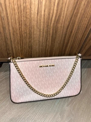 Michael Kors - Snygg ljusrosa väska från Michael Kors med guldfärgad kedja och dragkedja. Väskan har diskret MK-mönster över hela ytan och logga framtill. Insidan är fodrad med beige tyg och har små fack för kort. Perfekt för att lyfta din outfit.