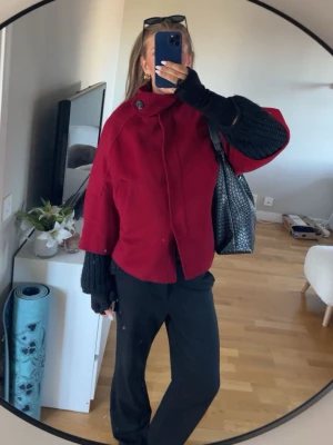 Röd poncho med knappdetalj - Snygg röd poncho/kappa med hög krage och stor knappdetalj vid halsen. Kappan har en lös och boxig passform med vida ärmar. Stilren och trendig design som sticker ut. Perfekt till våren