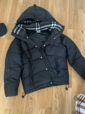 Svart pufferjacka med rutigt foder - Snygg svart pufferjacka med stor huva och rutigt foder i grått, vitt och rött. Jackan har dragkedja och tryckknappar framtill samt två dragkedjeförsedda fickor. Dragsko i nederkant för justerbar passform. Perfekt för kalla dagar.