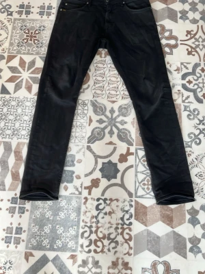 Svarta slim fit jeans från tiger of Sweden  - Säljer ett par svarta slim fit jeans med klassisk femficksdesign och raka ben. Jeansen har svarta sömmar och är tillverkade i ett mjukt denimtyg med lite stretch för extra komfort. Perfekta till en avslappnad streetwear-look.