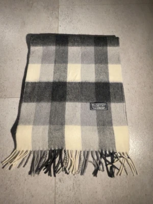 Rutig halsduk från Burberrys - Klassisk halsduk från Burberrys i rutigt mönster med nyanser av grått, svart och vitt. Tillverkad i 100% lammull och har fransar i båda ändar. Märkt med Burberrys of London, Made in England. Perfekt accessoar för kyliga dagar.