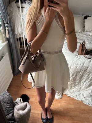 Vit kort studentklänning - Säljer en superfin vit kort klänning med spetsdetaljer vid midjan, halsen och nederkanten. Klänningen har en halterneck-modell och är ärmlös, vilket ger en fräsch och somrig look. Perfekt för dig som vill ha något stilrent och sött till sommaren!