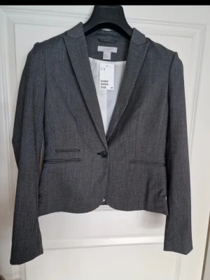 Ny grå kavaj från H&M med smal passform med etikett - Stilren grå kavaj från H&M med smal passform och klassisk krage. Kavajen har två fickor framtill och stängs med en knapp. Perfekt för en dressad look och enkel att matcha med olika outfits. Ger slank figur. 