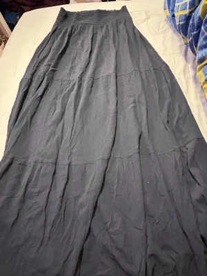 Blå långkjol med smock i midjan - En blå långkjol med smockad midja och lätt utställd passform. Kjolen har flera sömmar som ger en bohemisk känsla och är gjord i ett mjukt, tunt tyg som faller snyggt. Perfekt att styla med en enkel topp eller stickad tröja.