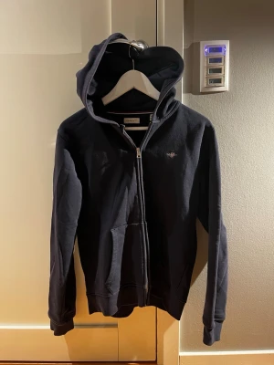 Mörkblå zip hoodie från GANT - Klassisk svart hoodie från GANT med dragkedja framtill och broderad logga på bröstet. Hoodien har huva med snörning och känguruficka. Tillverkad i mjukt bomullsmaterial som är skönt att ha på sig. Perfekt för en avslappnad och stilren look. 