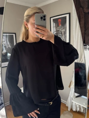 Svart blus med volangärmar - Svart blus med rund hals och breda volangärmar. Blusen har en loose passform och är tillverkad i ett mjukt, flowy material som känns skönt mot huden. Perfekt för dig som gillar stilrena och trendiga plagg.